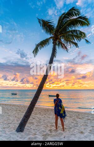 Vista posteriore della donna con sarong ammirando l'oceano al tramonto sulla spiaggia orlata di palme, le Morne Brabant, Black River, Mauritius, Oceano Indiano, Africa Foto Stock