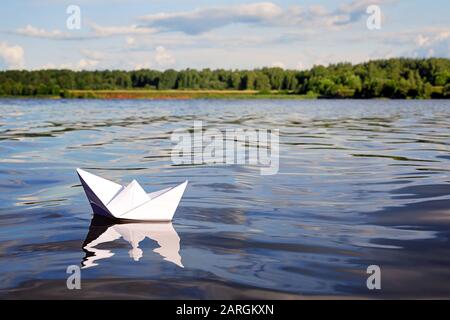 Piccola barca di carta che galleggia su acque blu calmo del fiume, foresta verde, cielo blu con le nuvole estive chiare sull'orizzonte. Libertà, avventura, sogno e tra Foto Stock