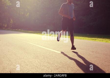 Uomo jogging nel parco estivo, silhouette di running guy in backlight casting ombra sul percorso asfalto. Foto Stock