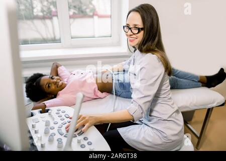 Giovane medico sorridente che muove il trasduttore a ultrasuoni sulla pancia africana donna e reni mentre guarda lo schermo in ospedale Foto Stock