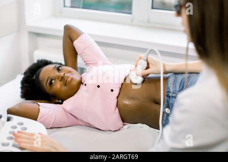 Dispositivo scanner a ultrasuoni nella mano di un medico professionista che esamina giovane donna incinta africana paziente, facendo ecografia addominale Foto Stock