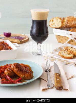 Il piatto con porzione di salsicce cotte in birra scura con cipolla rossa, paprika rossa e gialla, peperoncino rosso caldo, pane e glas al rosmarino ritorto Foto Stock