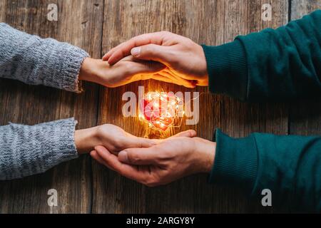 Due mani che tengono una forma di cuore rosso coperto con luci a LED su legno e bokeh luci sfondo. San Valentino e concetto romantico Foto Stock