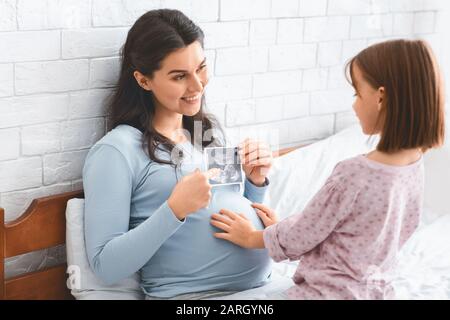 Donna incinta allegra che mostra la scansione ecografica a sua figlia piccola Foto Stock