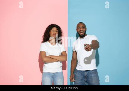 Usando il telecomando della TV, è arrabbiata. Giovane uomo e donna in abiti casual su sfondo rosa, blu bicolored. Concetto di emozioni umane, expation facciale, relazioni, annuncio. Bella coppia afro-americana. Foto Stock