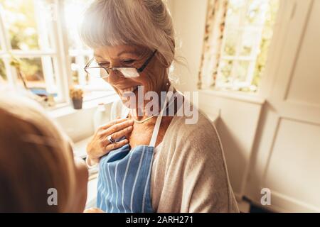 Donna sorridente in piedi nella sua casa in un grembiule. Felice vecchia donna in piedi in cucina e sorridente. Foto Stock