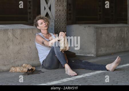 una donna punk sotto un ponte con bottiglie di alcol nelle vicinanze e una bottiglia in un sacchetto di carta marrone in mano Foto Stock