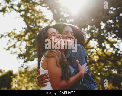 Ritratto sorridente di una giovane femmina diversa con gli occhi chiusi coccolarsi e amarsi l'un l'altro nel parco con luce solare che splende attraverso Foto Stock