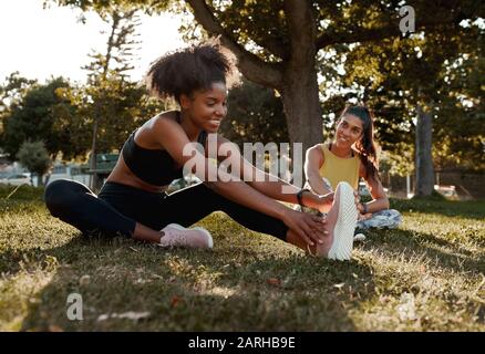 Sorridendo due diverse sportive fit giovani amici femminile tendendo le gambe nel parco - afroamericano nero femmina riscaldamento con la sua amica Foto Stock