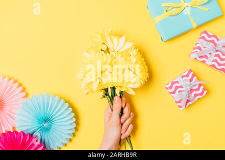 Mani di donna che tengono bouquet di fiori accanto a scatole regalo e decorazioni per le vacanze su sfondo giallo brillante, piatto lay. Foto Stock