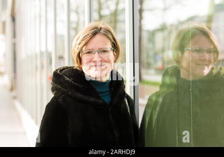 Ritratto di una donna di trent'anni con un cappotto invernale nero, in piedi rilassato contro un moderno edificio d'ufficio, che riflette nel vetro Foto Stock