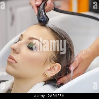 Bella giovane donna lava i capelli al parrucchiere. Le mani del parrucchiere sostengono la testa, gli occhi sono chiusi, l'acqua scorre giù i capelli Foto Stock