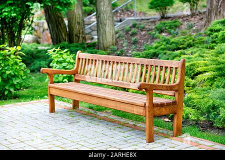 Una bella ed elegante panchina in rovere artigianale sorge in un parco in un'area ricreativa su lastre di pavimentazione con cespugli verdi e grandi alberi sul retro. Foto Stock