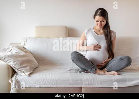 Giovane donna incinta seduta su divano bianco e tenendo le mani sul ventre Foto Stock
