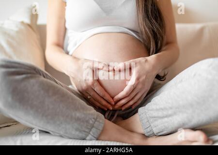 Giovane donna incinta seduta su divano bianco e tenendo le mani sul ventre Foto Stock
