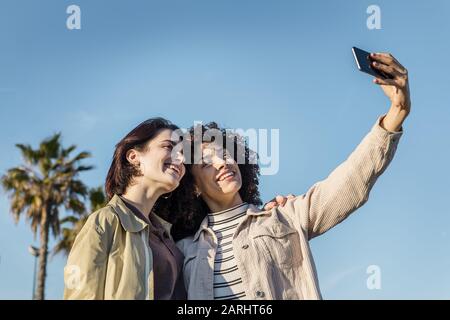 giovane coppia interracial di donne che fanno una foto selfie in uno sfondo blu cielo con un telefono cellulare, concetto di amicizia femminile e diversità razziale Foto Stock