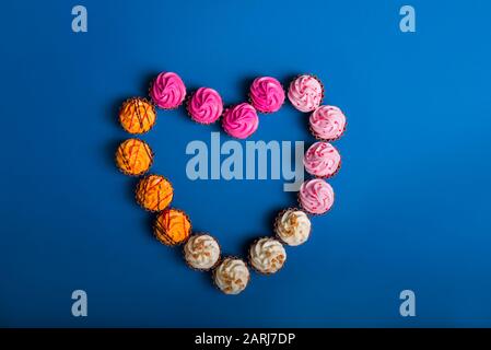 Top view Allsmista dolci al cioccolato con crema multicolore ripieno disposto a forma di cuore su sfondo classico di colore blu. Buon San Valentino. Lo Foto Stock