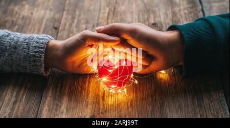 Due mani che tengono una forma di cuore rosso coperto con luci a LED su legno e bokeh luci sfondo. San Valentino e concetto romantico Foto Stock