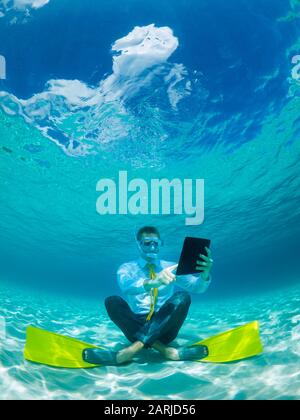 Snorkeling uomo d'affari in camicia e cravatta e pinne corrispondenti utilizzando un computer tablet seduto sott'acqua in acque tropicali turchese mare Foto Stock