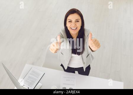 Vista dall'alto. Positivo fiducioso business woman sorride ha sollevato il pollice in su, mentre in piedi al tavolo sul posto di lavoro in ufficio. Foto Stock