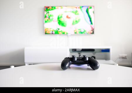 Una vista di un controller per videogiochi nero senza logo su un tavolo bianco. La TV è sullo sfondo. Foto Stock