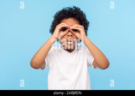 Curioso bambino esplorare il mondo. Ritratto di noia curioso ragazzino curly guardare attraverso le dita a forma di binocolo ed esprimere stupore. Foto Stock