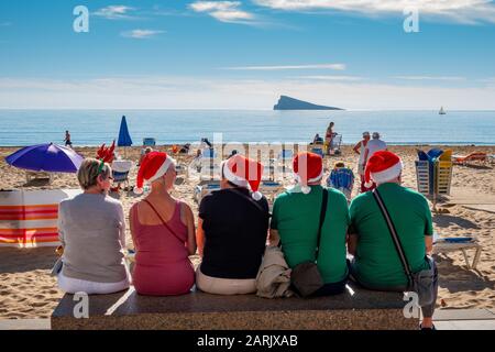 Gruppo di mezza età che indossa cappelli rossi santa seduti su una panchina di fronte alla spiaggia e al mare in inverno, in una luminosa giornata di sole a Benidorm, Spagna. Due uomini e t Foto Stock