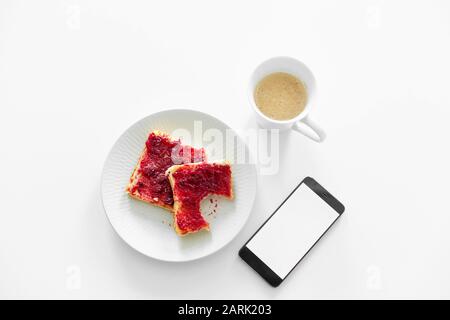 La colazione è a tavola. Una tazza di caffè e toast con marmellata di ciliegie accanto al telefono. Foto Stock