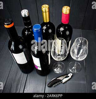 Bottiglie di vino rosso di produttori diversi con due bicchieri di vino frizzante e un cavatappi per la degustazione, in uno spazio di legno grigio ben illuminato Foto Stock