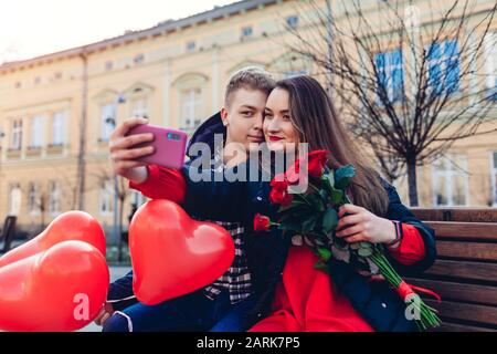 San Valentino. Coppia in amore prendere selfie utilizzando il telefono con bouquet di fiori di rose in data in città. Giorno donne Foto Stock