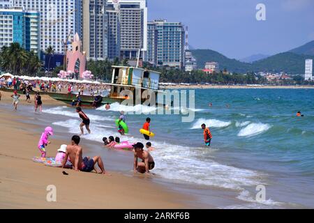 La gente gode il lungomare e la spiaggia nella località balneare di Nha Trang in Vietnam, Sud Est asiatico, Indochina, Asia, in una giornata calda e soleggiata. Per quanto riguarda la chiatta naufragata in background: Nel dicembre 2019 una chiatta del traghetto che porta il numero KH-98668-TS ha deviato ed è stato lavato nella spiaggia di Nha Trang ed è ancora bloccata sulla sabbia ad est di Tran Phu Street, Nha Trang, il 29 gennaio 2020. Foto Stock
