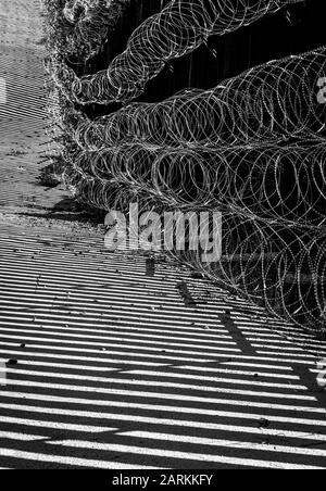Vista astratta della recinzione di confine internazionale USA/Messico con filo di rasoio con luce e ombre drammatiche a Nogales, AZ, USA, in bianco e nero Foto Stock
