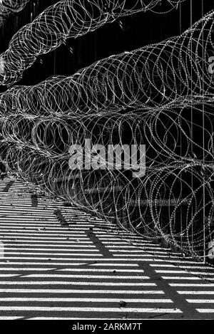 Vista astratta della recinzione di confine internazionale USA/Messico con filo di rasoio con luce e ombre drammatiche a Nogales, AZ, USA, in bianco e nero Foto Stock