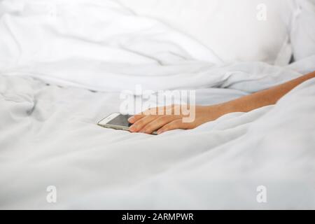 La mano da vicino tiene il telefono a letto. La ragazza si è addormentato con un telefono in mano ed è addicted. Oppure una persona si rivolge a un telefono cellulare per disattivare la sveglia. Foto Stock