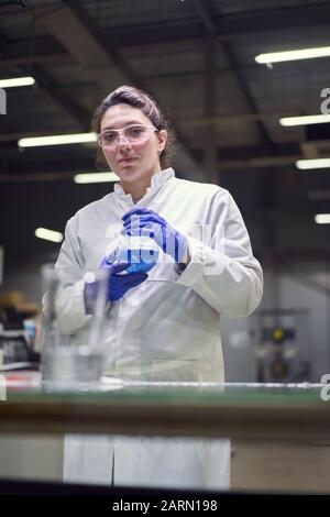 Donna da laboratorio brunetta che guarda la macchina fotografica in occhiali e cappotto bianco con matraccio sperimentale con liquido blu nelle sue mani conduce esperimenti su de Foto Stock