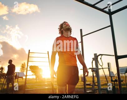 Felice uomo giovane che si gode ascoltare la musica sulle cuffie dopo l'esercizio nel parco all'aperto palestra in estate giornata di sole Foto Stock