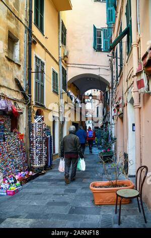 Vicolo stretto ('carruggio') nel centro storico di Sanremo, quartiere Pigna, con un uomo che porta borse per lo shopping e un negozio turistico, Imperia, Liguria Foto Stock