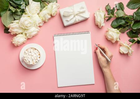 Le mani femminili scrivono in taccuino e bouquet rose bianche intorno sul tavolo rosa. Vista dall'alto con spazio di copia. Foto Stock