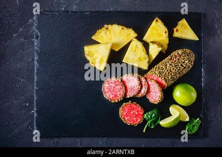 salame rivestito con pepe verde intero e affettato su una tavola di pietra nera e servito con ananas e lime, piatto, primo piano, orizzontale Foto Stock