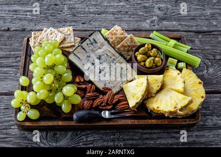 Formaggio blu servito con pecan, uva verde, sedano bastoni, biscotti cracker, fette di ananas e olive su un vassoio di legno rude su un tavolo rustico, hor Foto Stock