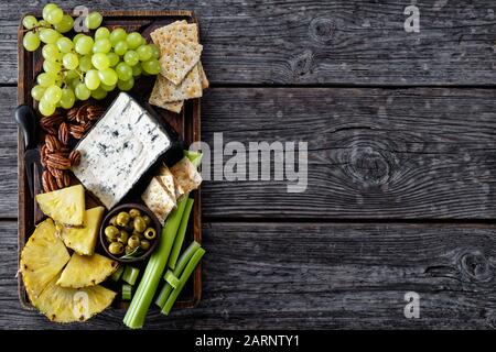 Formaggio blu servito con pecan, uva verde, sedano bastoni, biscotti cracker, fette di ananas e olive su un vassoio di legno rude su un tavolo rustico, hor Foto Stock