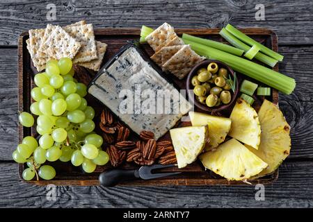 Formaggio blu servito con noci pecan, uva verde, sedano bastoni, biscotti cracker, fette di ananas e olive verdi su un vassoio di legno rude su un rustico Foto Stock