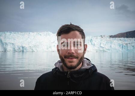 Felice e sorridente giovane uomo che si trova di fronte al ghiacciaio Eqip Sermia chiamato ghiacciaio Eqi. Turistico con enorme parete di ghiaccio sullo sfondo. Il concetto di Foto Stock