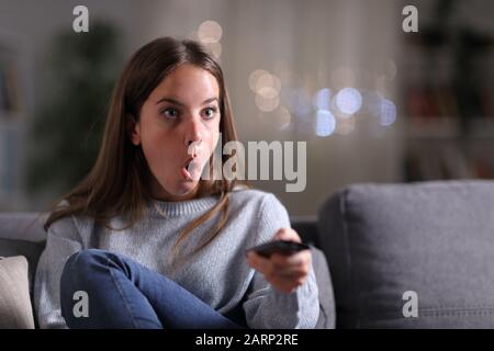 Sorpresa donna guardare la tv seduto su un divano a casa di notte Foto Stock