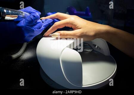 primo piano per rimuovere lo smalto gel dai chiodi di una mano femmina con un apparecchio speciale. Le mani femminili nei guanti blu tengono una macchina per manicure. Foto nella d Foto Stock