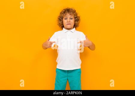 Ragazzino con i capelli ricci in colorate t-shirt e shorts mostra i pollici per se stesso isolato su sfondo giallo. Foto Stock