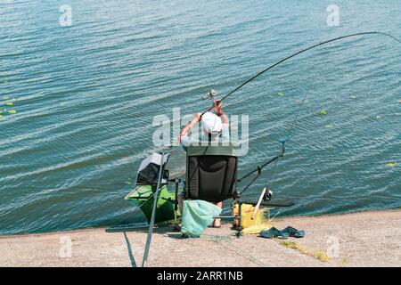 Uomo adulto anziano sulla riva del fiume in Ucraina che getta il fishrod. Fishman è seduta sulla sedia circondata da attrezzature da pesca. Immagine a colori con c Foto Stock