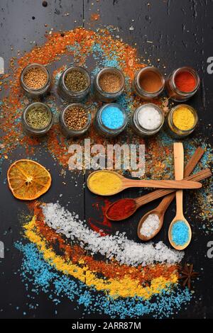 Cucchiai di legno con paprika, curcuma e sale marino vicino agrumi e spezie mix su tavolo. Concetto di arte alimentare. Set di spezie su sfondo grigio scuro. Composizione di sale colorato e spezie in prossimità di vasi con erbe Foto Stock