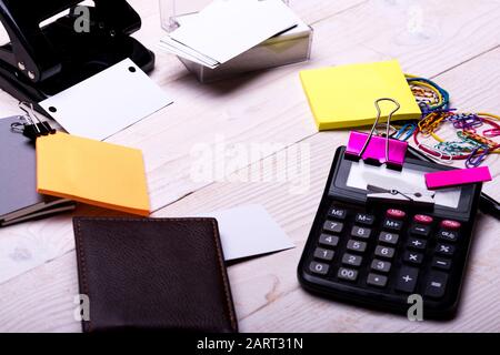 Cancelleria e calcolatrice. Concetto di accessori e idee per il business. Puncher, portafoglio e notebook su sfondo bianco in legno. Biglietti da visita nel supporto, raccoglitori, fermagli ed elastici vicino alle note adesive Foto Stock