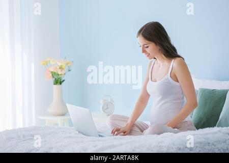 Bella donna incinta con un computer portatile seduto sul letto Foto Stock